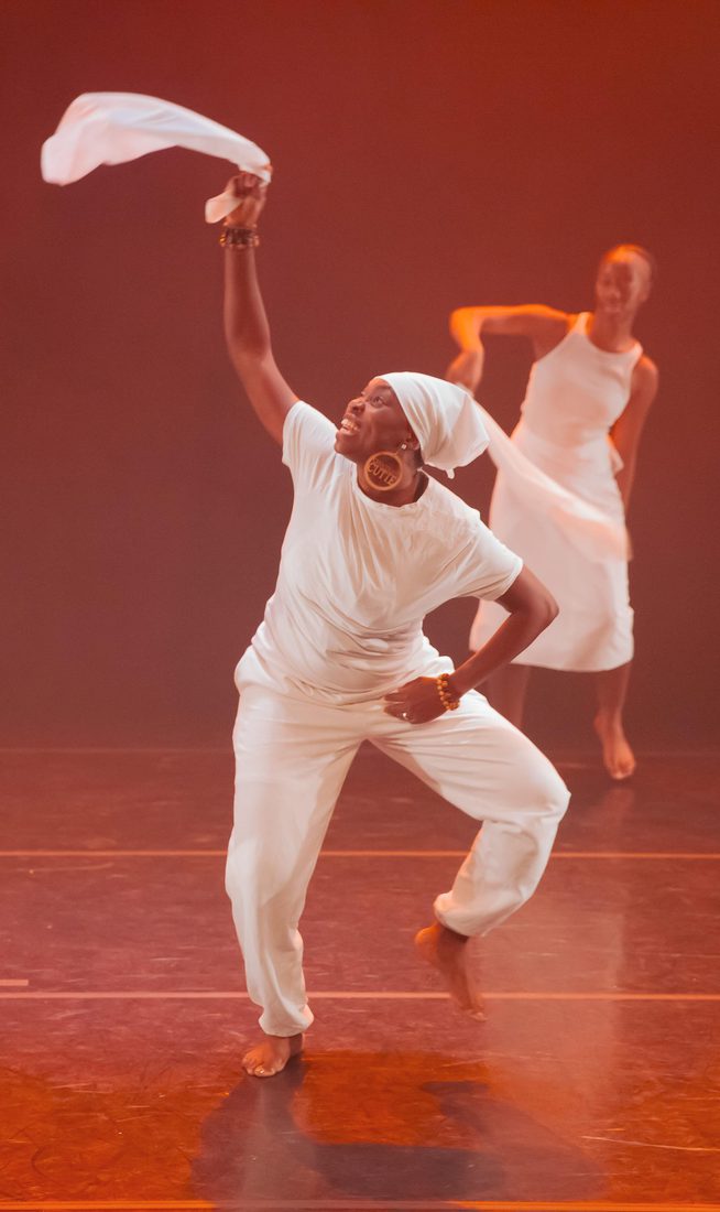 Four dancers perform on a stage lit with warm orange light. Two dancers in the foreground move with one arm raised, each holding a piece of white fabric, while two others are visible in the background. All are dressed in white, and several appear to be mid-step or airborne.
