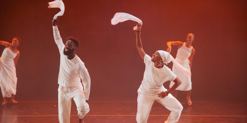 Four dancers perform on a stage lit with warm orange light. Two dancers in the foreground move with one arm raised, each holding a piece of white fabric, while two others are visible in the background. All are dressed in white, and several appear to be mid-step or airborne.