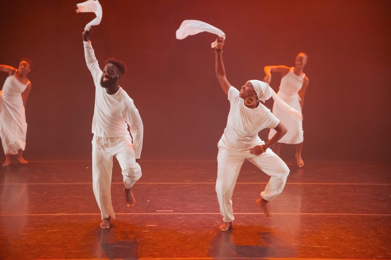 Four dancers perform on a stage lit with warm orange light. Two dancers in the foreground move with one arm raised, each holding a piece of white fabric, while two others are visible in the background. All are dressed in white, and several appear to be mid-step or airborne.