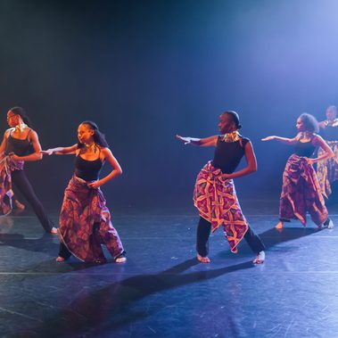 Five dancers move across the stage with one arm extended forward and the other held at the waist. They wear black tops, patterned wrap skirts, and large metallic neckpieces. Blue and amber stage lighting illuminates the group against a dark background.