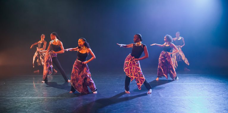 Five dancers move across the stage with one arm extended forward and the other held at the waist. They wear black tops, patterned wrap skirts, and large metallic neckpieces. Blue and amber stage lighting illuminates the group against a dark background.