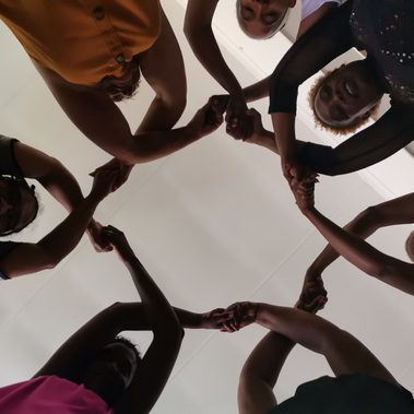 Looking up from inside a circle of seven women whose hands are lifted above their heads, crossed and linked to make repetitive, connected pattern.