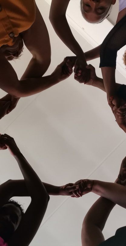 Looking up from inside a circle of seven women whose hands are lifted above their heads, crossed and linked to make repetitive, connected pattern.