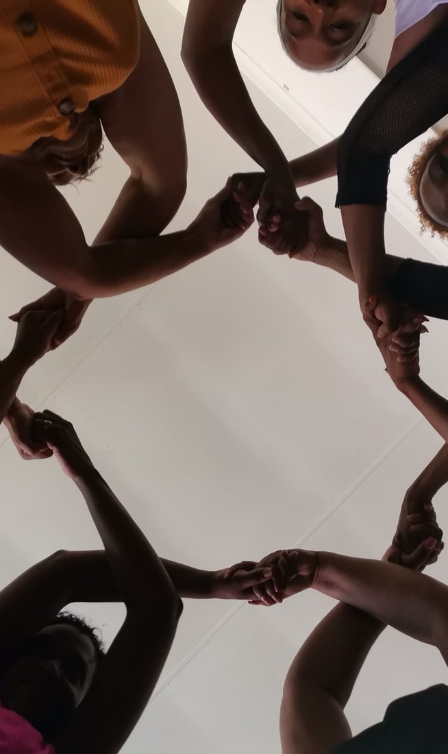 Looking up from inside a circle of seven women whose hands are lifted above their heads, crossed and linked to make repetitive, connected pattern.