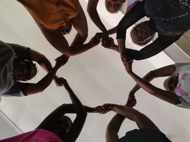 Looking up from inside a circle of seven women whose hands are lifted above their heads, crossed and linked to make repetitive, connected pattern.