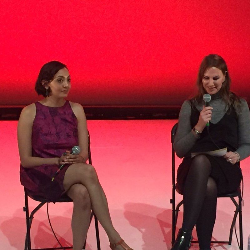 Two people sit on chairs onstage against a red backdrop, with Suzanne on the right. The person on the left, holding a microphone in her lap, wears a sleeveless purple dress. The person on the right holds a microphone and reads from a paper, dressed in a grey turtleneck with a black dress layered over it. A bottle of water sits on the floor beside them.