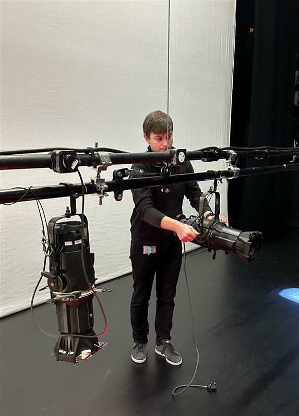 Luke on stage adjusting a reflector spotlight. He has short brown hair and is wearing a black long-sleeve top, black trousers, and grey sneakers.