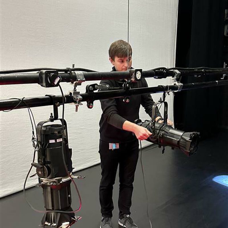 Luke on stage adjusting a reflector spotlight. He has short brown hair and is wearing a black long-sleeve top, black trousers, and grey sneakers.