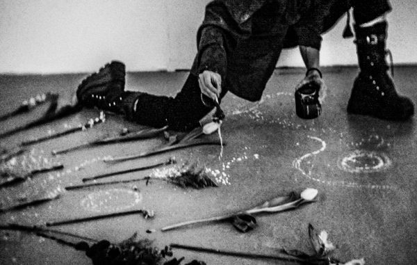 A black-and-white scene shows a person kneeling on the floor, pouring or drawing a swirling line of white granules from a small container. Long-stemmed flowers are arranged in rows across the floor around them, creating a ritual-like or artistic installation.