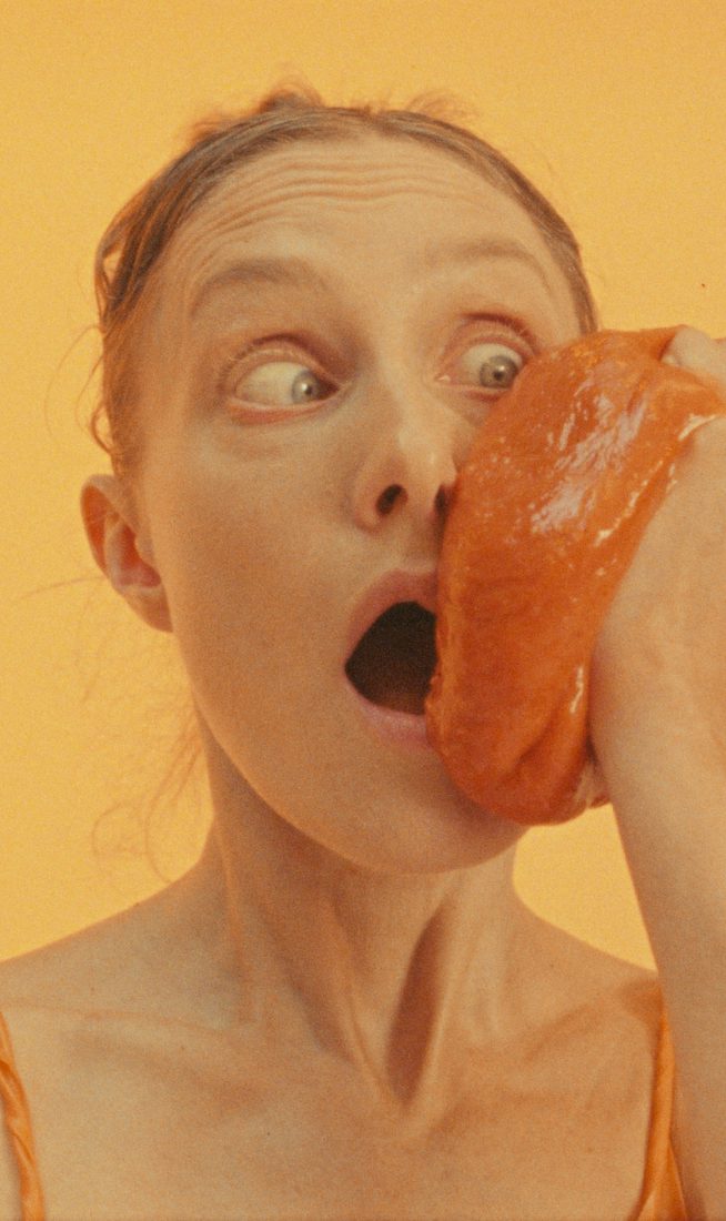 A woman against an orange background wearing an orange costume holding an orange jelly to her face.