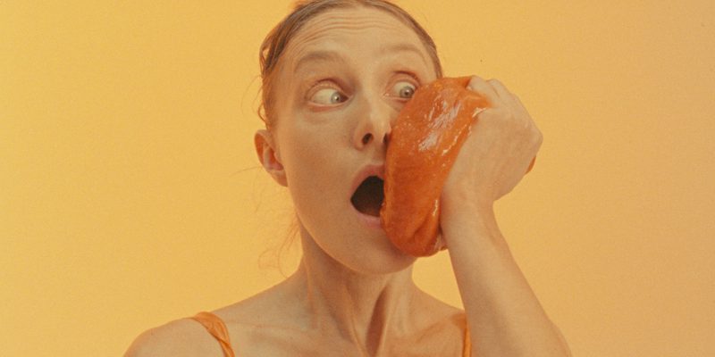 A woman against an orange background wearing an orange costume holding an orange jelly to her face.