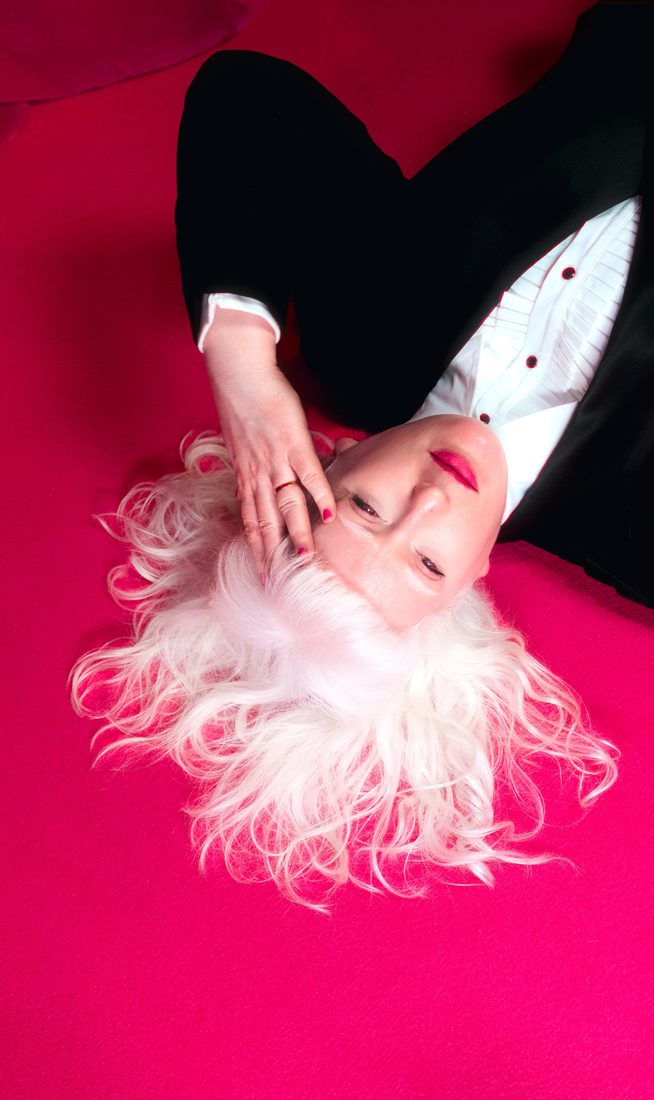 A photograph of Jo Bannon lying on a vibrant pink carpet with her white hair fanned out around her head. She is wearing a black suit and white shirt and hot pink lipstick. Her hand is held to her forehead. A small white rabbit is positioned nearby. The background features a rich pink curtain.