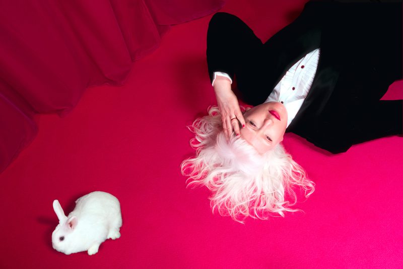 A photograph of Jo Bannon lying on a vibrant pink carpet with her white hair fanned out around her head. She is wearing a black suit and white shirt and hot pink lipstick. Her hand is held to her forehead. A small white rabbit is positioned nearby. The background features a rich pink curtain.