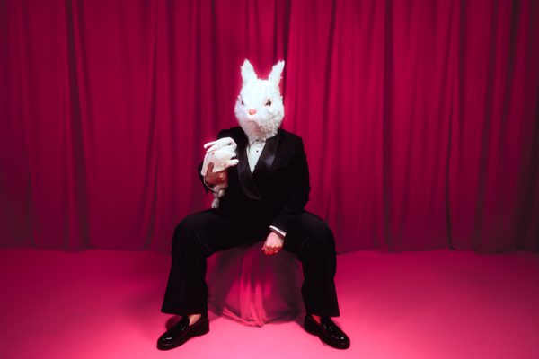 A photograph of Jo Bannon wearing a white rabbit mask, seated in a commanding pose on a pink box placed on a vibrant pink carpet, with her legs apart. She is dressed in a black trouser suit and white shirt. She is holding a live white rabbit in one arm. The backdrop features bold pink velvet drapes.
