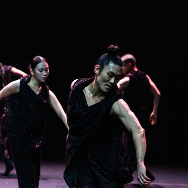 A group of contemporary dancers in black, sequined sleeveless costumes perform on a dark stage under pink and purple lighting. In focus, a dancer leans forward with an extended arm, others around them mirroring the pose.