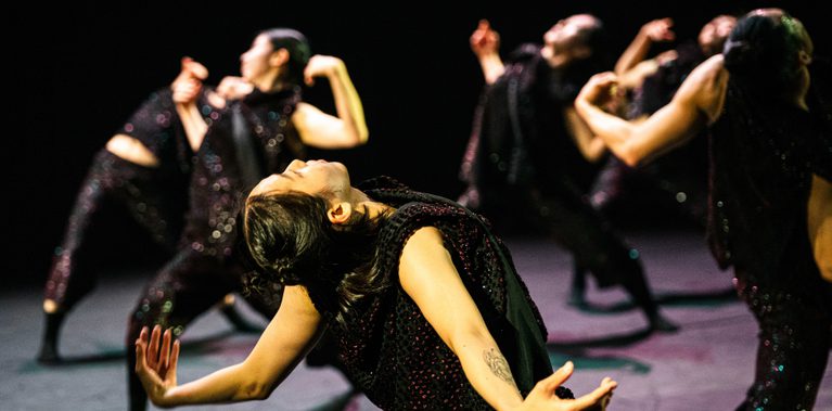 On stage, a group of dancers wearing black sequined sleeveless tops and matching trousers perform in a loose circular formation. One dancer is in sharp focus in the foreground. All balance on one leg while arching slightly back, with their arms opened out to the sides.