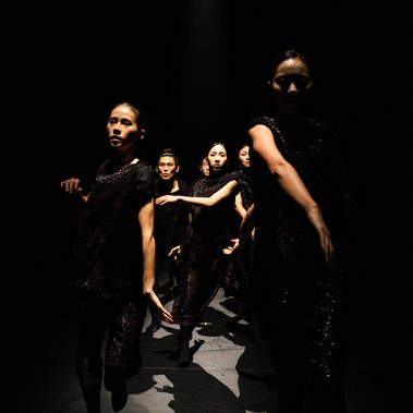 A group of dancers performs on a stage. They wear draped black sequined tops and matching trousers that catch the light subtly.