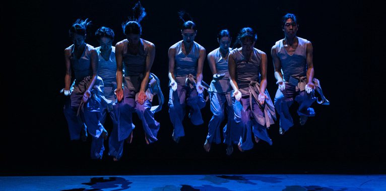 On a dark blue-lit stage, a group of dancers in matching light sleeveless tops and loose trousers is caught mid-jump in unison, all reaching a similar height. Their arms are extended forward along their bodies, hands turned toward the audience with palms open. Their heads tilt downward, knees bent, and beneath them, pronounced shadows of their silhouettes stretch across the stage.