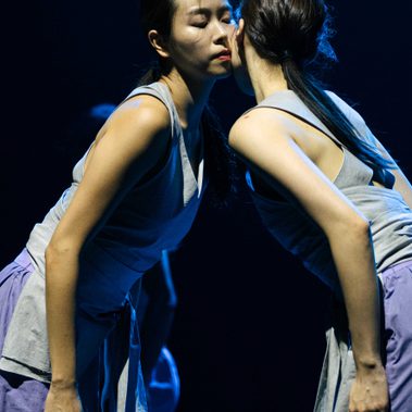 Two people, wearing matching costumes with light sleeveless tops featuring apron-like bottoms and long loose ties, paired with loose light purple trousers, are captured from the knees up. They face each other, leaning in so their left cheeks touch, with their arms extended alongside their bodies.