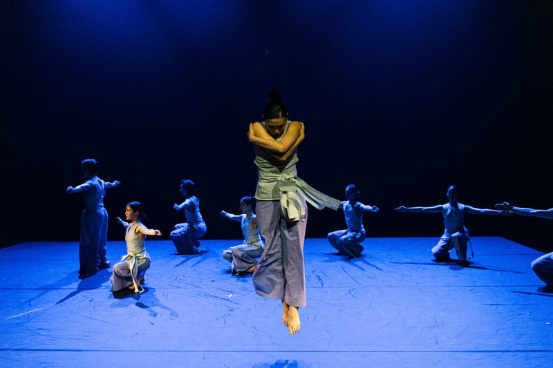 A group of dancers in matching costumes of light, draped sleeveless tops and loose light purple trousers is spread across a blue-lit stage. In the forefront, a dancer crouches, arms crossed over their chest in a self-contained, inward gesture, head tilted downward. Behind them, the ensemble holds a static pose, crouching with arms extended to the sides, while one dancer stands sideways, likewise stretching their arms outward.