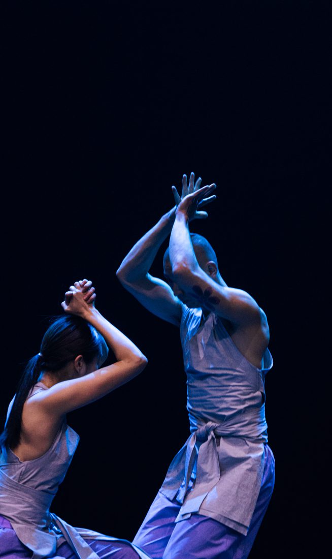 Two dancers, wearing matching light, draped sleeveless tops and loose light purple trousers, face each other. Both raise their bent arms, joining their hands above their heads. One crouches, lowering themselves by bending their knees, while the other remains standing, creating a sense of height contrast between them.
