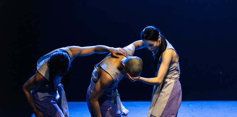 Three performers on a blue-lit stage, wearing matching costumes consisting of light grey sleeveless tops and loose light purple bottoms, captured from the knees up. From left to right: the first two performers are aligned, facing the same direction. Both lean their torsos to the right, bow their heads, bend their knees, and extend their left arms forward to rest them on the shoulder of the performer in front. On the far right, the third performer faces the other two, supporting the middle performer’s head with their hand.