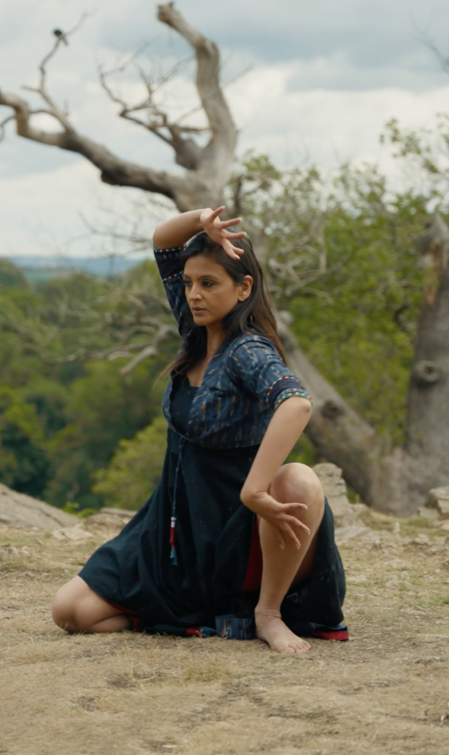 A woman in a dark blue and black dress kneels barefoot on dry, rocky ground, holding a poised classical dance position with one arm curved above her head and the other bent near her knee. Behind her stretches a wide landscape of green hills and trees, under a dramatic sky. A gnarled, leafless tree adds striking contrast in the background.