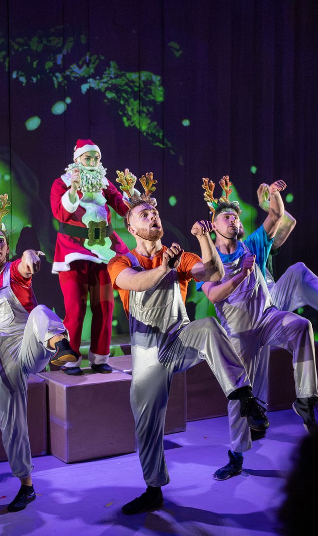Santa guides 5 performers that are wearing reindeer antlers as they dance together in formation. Santa stands on cardboard boxes. The background shows an abstract projected image of green blobs.