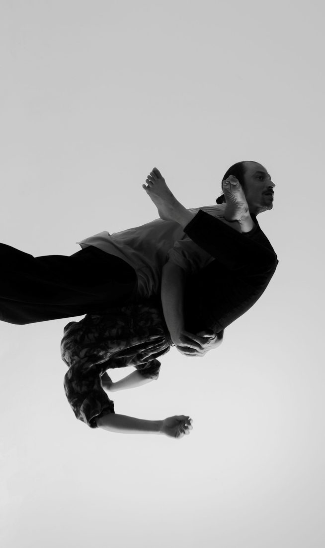 In a black-and-white image, two performers are captured in a partnered lift, with one suspended upside down over the other's shoulder.