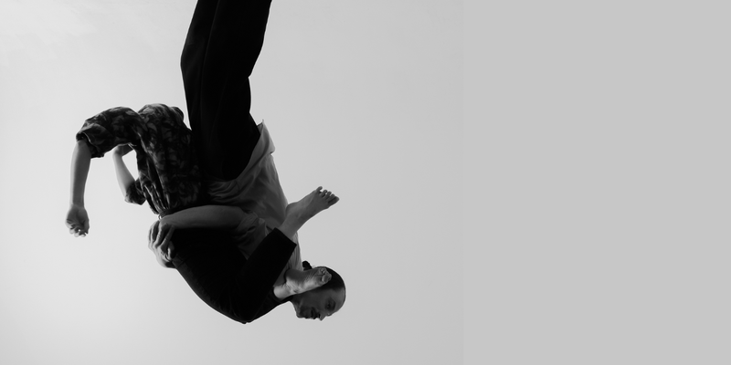 In a black-and-white image, two performers are captured in a partnered lift, with one suspended upside down over the other's shoulder.