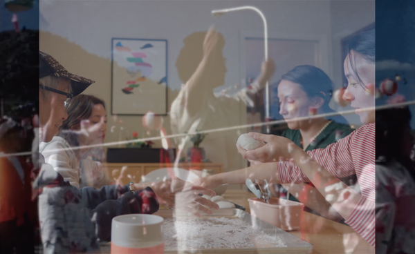 A double-exposure image layers a group of people shaping dough around a table with a translucent figure standing and moving in the background. The overlapping scenes create a dreamlike effect, blending hands-on activity, conversation, and motion within a bright indoor room.