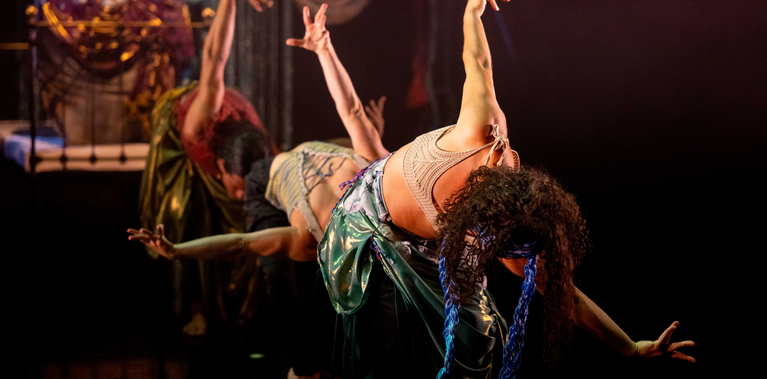 Three performers are on a dark stage, leaning back in a curtsy position with their arms raised. The central performer, in a shiny green skirt and a light top, is the clearest, while the others are slightly blurred in the foreground, creating a synchronized, fluid motion
