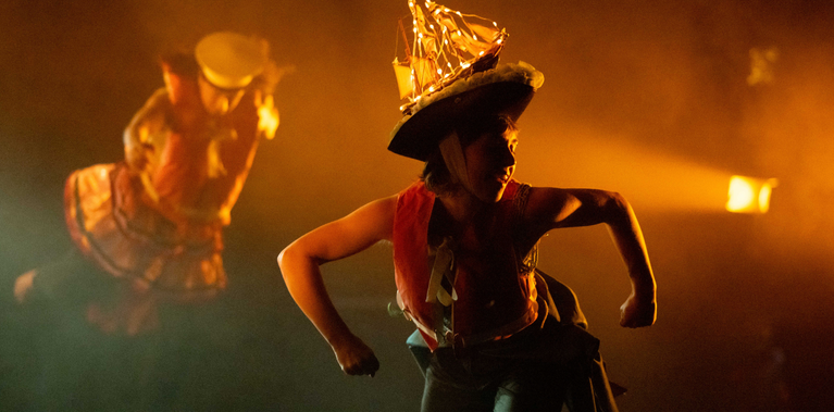 A performer in a captain's hat with a lit-up model ship on top crouches forward on a dimly lit, foggy stage. The scene is cast in an orange glow from the stage lights.