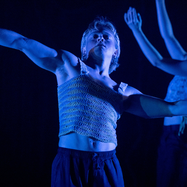 Three performers are in a choreographed pose under a vibrant blue light. The central performer extends their arms wide, with the other two performers in mirrored poses on either side, all wearing knit tops.
