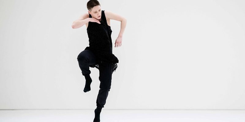 A dancer dressed in black with short dark hair is in the frame. The background is a white cream colour. The dancer jumps into the air, their right leg slightly raised as if they are running in the air. Their right arm is bent inwards towards the chest so the front of the hand touches their chin. The other arm is bent as well but is more to the side.