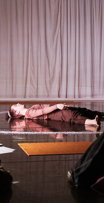 A person in the background lays on the black floor with a white ring of salt around their body and a spotlight on them, they are wearing burgundy clothes and their are two people watching them in the foreground