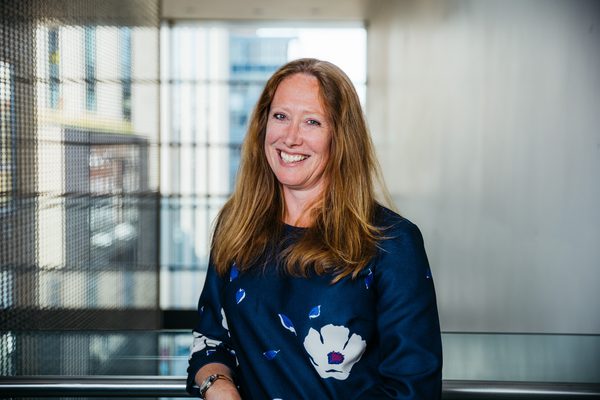 A woman with light red hair and a big smile looks at the camera. She is leaning on the metal railing and wearing a blue silk long sleeved top