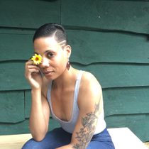 A woman with a grey strap top on covers her right eye with a yellow flower, she has tattoos on her left arm and short black hair