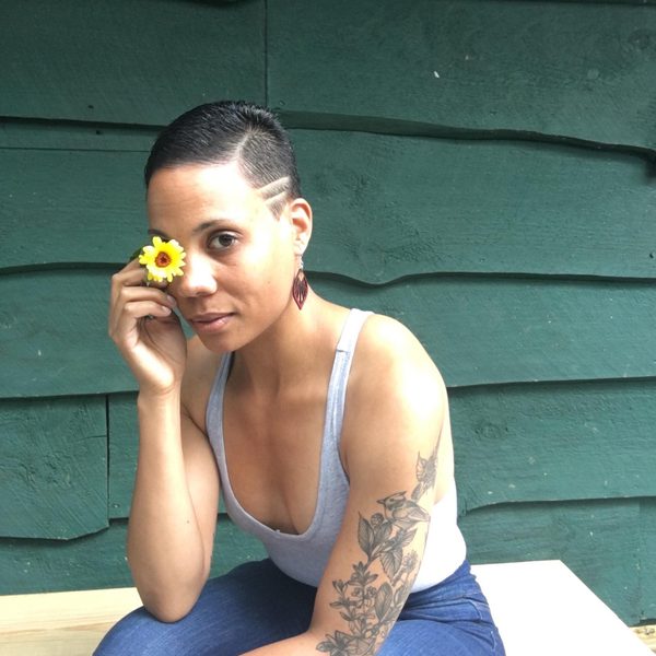 A woman with a grey strap top on covers her right eye with a yellow flower, she has tattoos on her left arm and short black hair