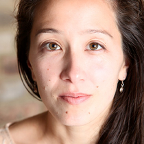 A woman with brown eyes looks directly into the camera, she is wearing a cream top and had brown long hair