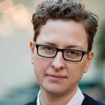 A woman with short hair, a soft smile and glasses looking directly into the camera, the background is blurry and her face is in focus.