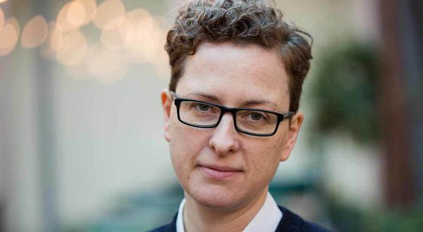 A woman with short hair, a soft smile and glasses looking directly into the camera, the background is blurry and her face is in focus.