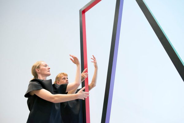 Two dancers hold their arms in curved positions wearing black. The background is white and there is a red, purple and green linear structure that surrounds them