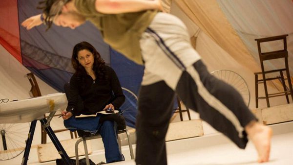 A woman sits on a chair with a notebook, she is watching a dancer in during a rehearsal, she has an intense look on her face