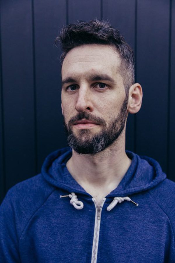 A man with short black hair and a beard is wearing a blue zip up hoodies and is looking into the camera with a serious expression