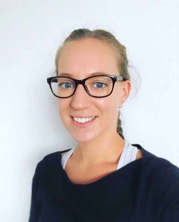 A woman with blonde hair, wearing a black long sleeved top is facing side on and smiling, she has black rimmed glassed and a white sports bra on