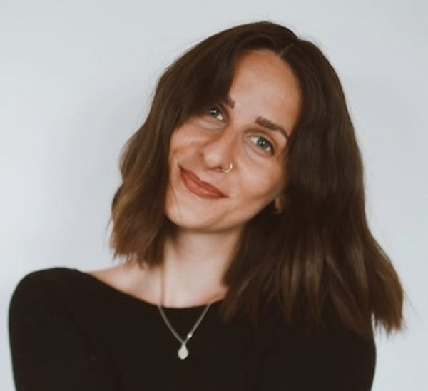 A woman with shoulder length wavy hair has her head tilted slightly to the right and she is smiling at the camera. She wears a black long sleeved top with a silver chain and pendant