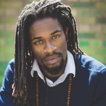 A man wearing a blue jumper, white shirt and a long beaded necklace looks directly into the camera