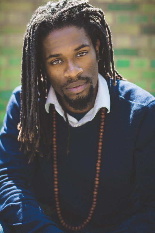 A man wearing a blue jumper, white shirt and a long beaded necklace looks directly into the camera
