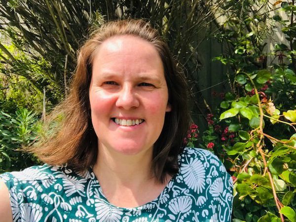 A woman with mid length brown hair is smiling and looking at the camera. She is wearing a green and white floral top and behind her there are plants and flowers.